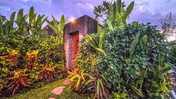 A small, rustic building with a red door, surrounded by lush tropical plants and a stone wall.