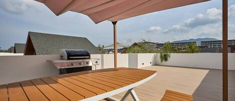 Un patio exterior con una mesa de madera y una parrilla, un paraguas grande y vistas a edificios y montañas.