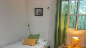 Une petite pièce bien rangée, avec un lit simple, une table de chevet avec une lampe, une fenêtre avec des rideaux et une prise électrique fixée au mur.