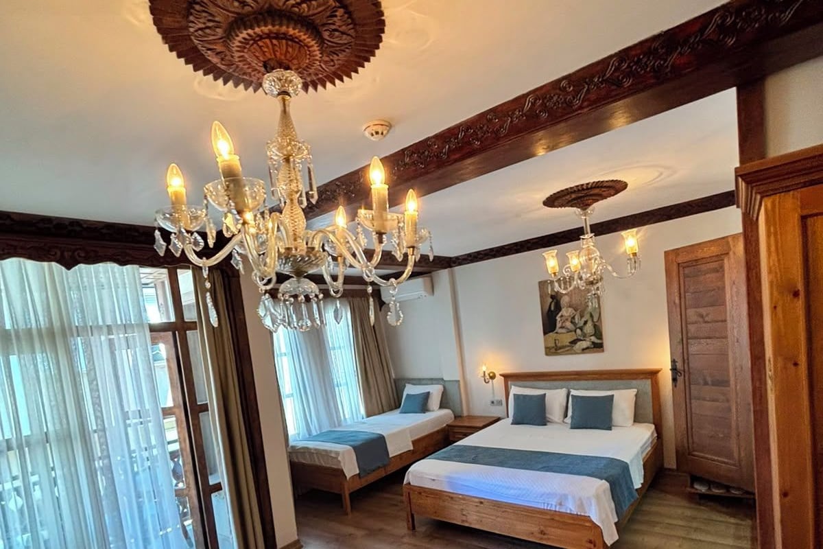 A hotel room with a chandelier, two beds, a wooden table, and a view through a window with sheer curtains.