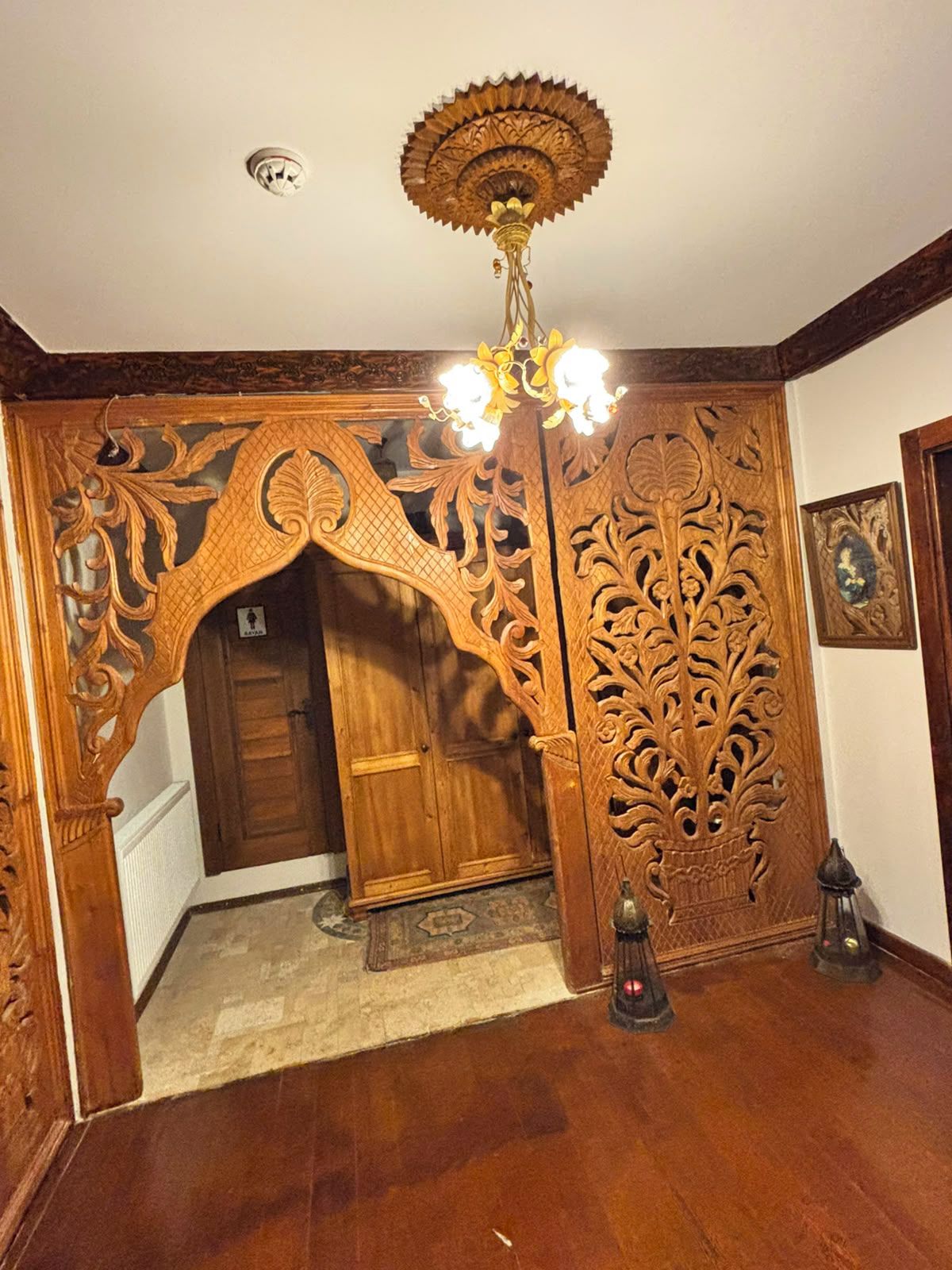 An ornate wooden doorway with intricate carvings, a chandelier, and a framed picture on the wall.