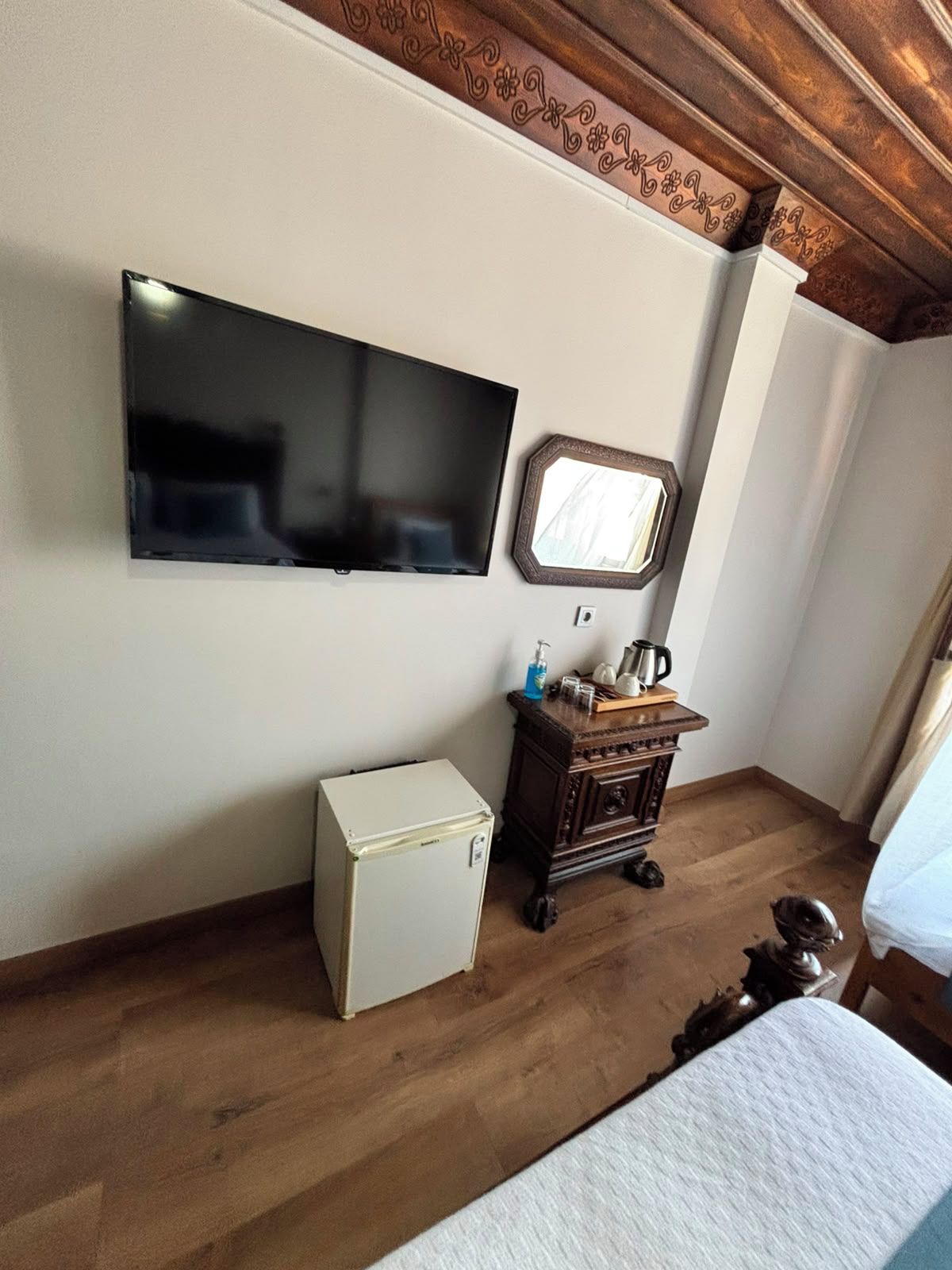 A hotel room with a flat-screen TV mounted on the wall, a small refrigerator, a wooden dresser with a mirror, and a bed with a white bedspread.