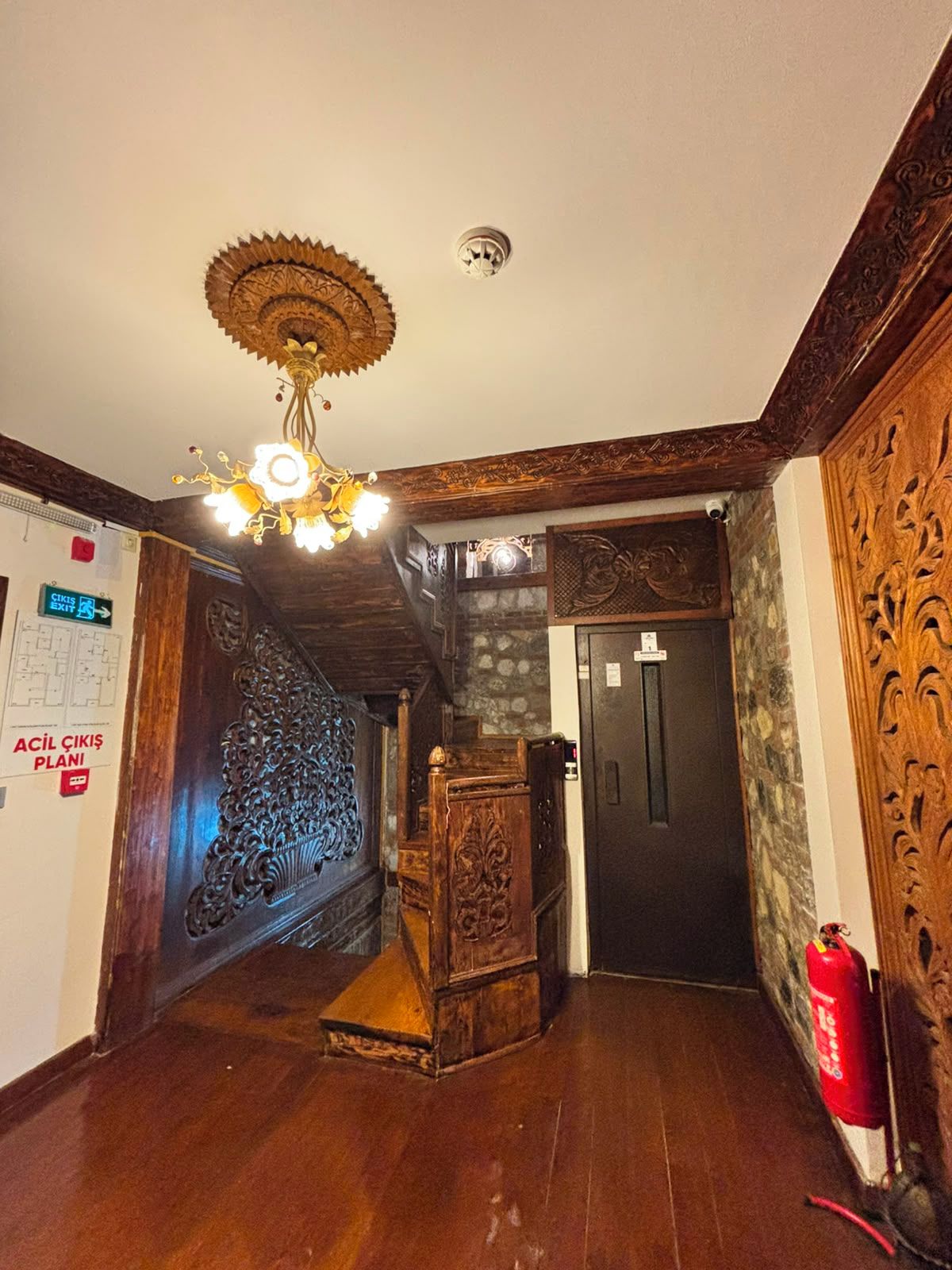A hallway with a wooden floor, a decorative ceiling light, a fire extinguisher, and a door with a sign above it.