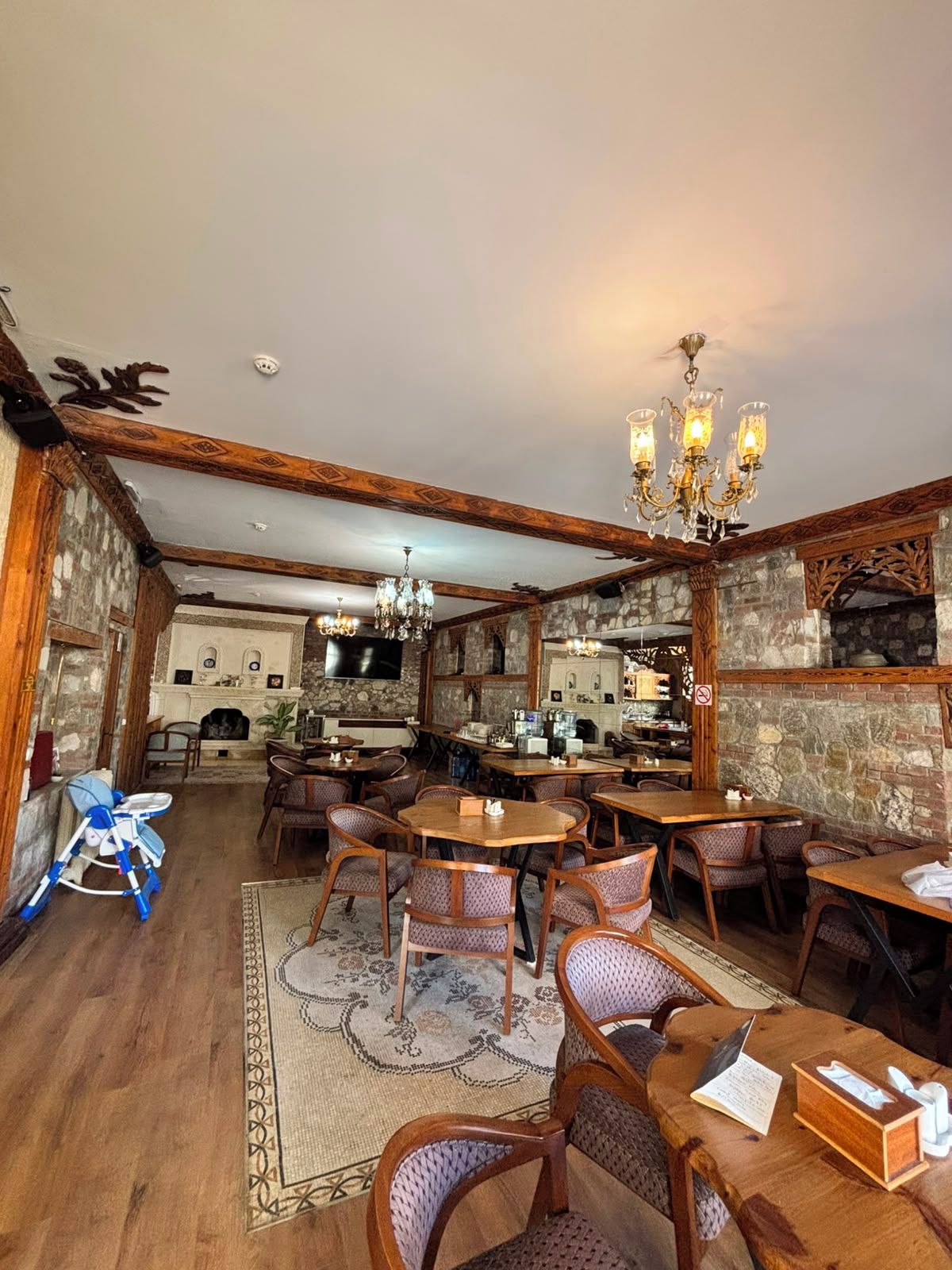 A restaurant with wooden beams, stone walls, and a chandelier. Tables and chairs are arranged for dining.