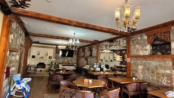 A restaurant with wooden beams, stone walls, and a chandelier. Tables and chairs are arranged for dining.