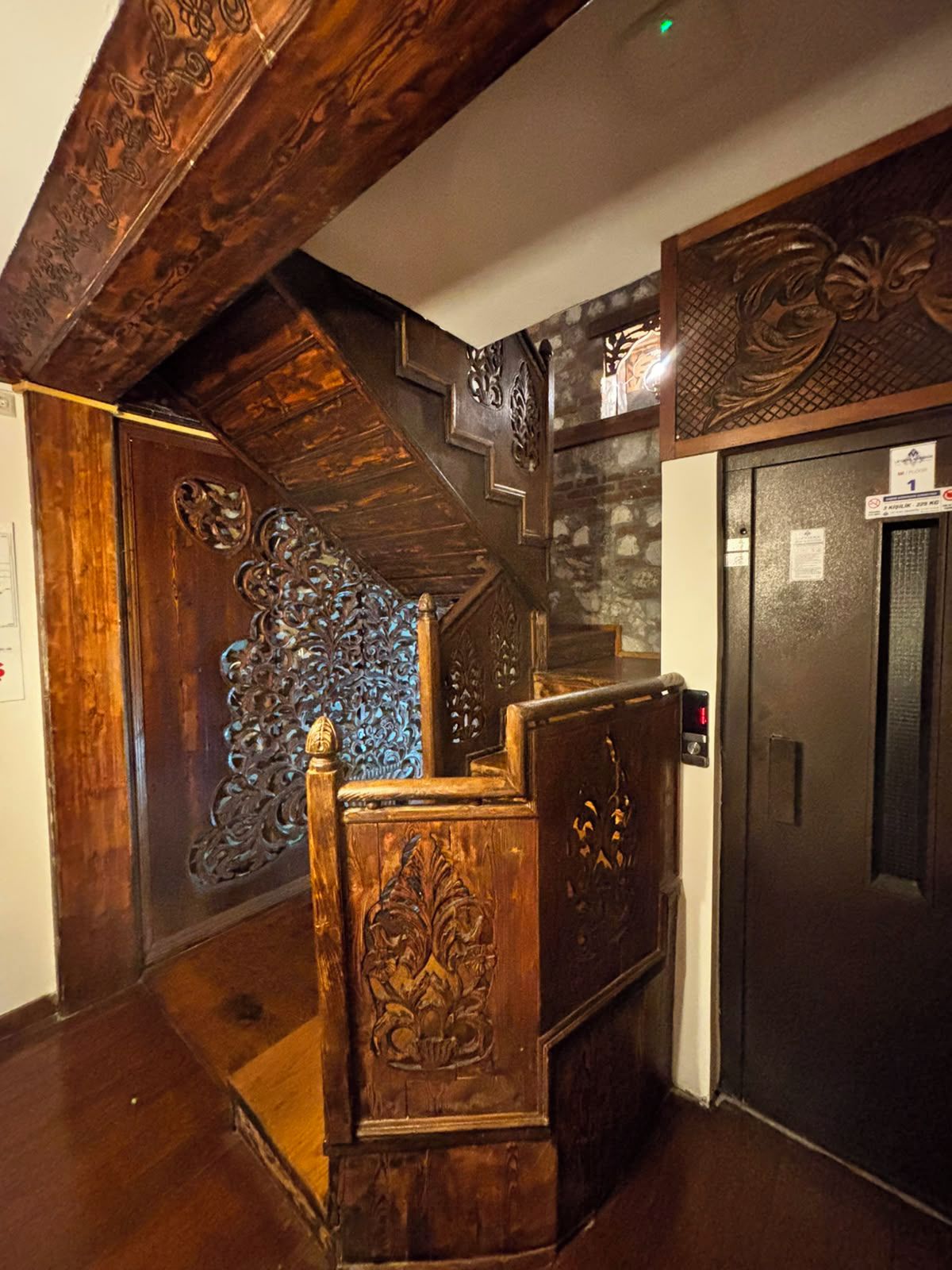 Una scala in legno con elaborate decorazioni intagliate, un ascensore scuro e un pannello decorativo a parete.
