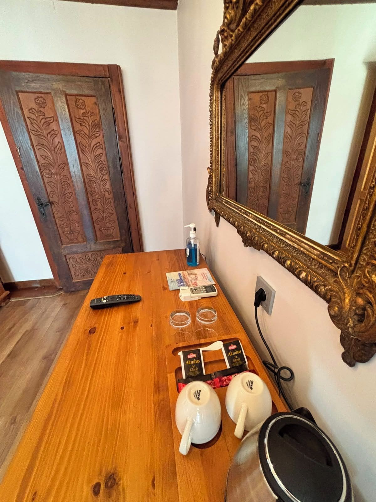A wooden table with a remote control, a bottle of hand sanitizer, two glasses, two white cups, and a black object on a wall with a mirror.