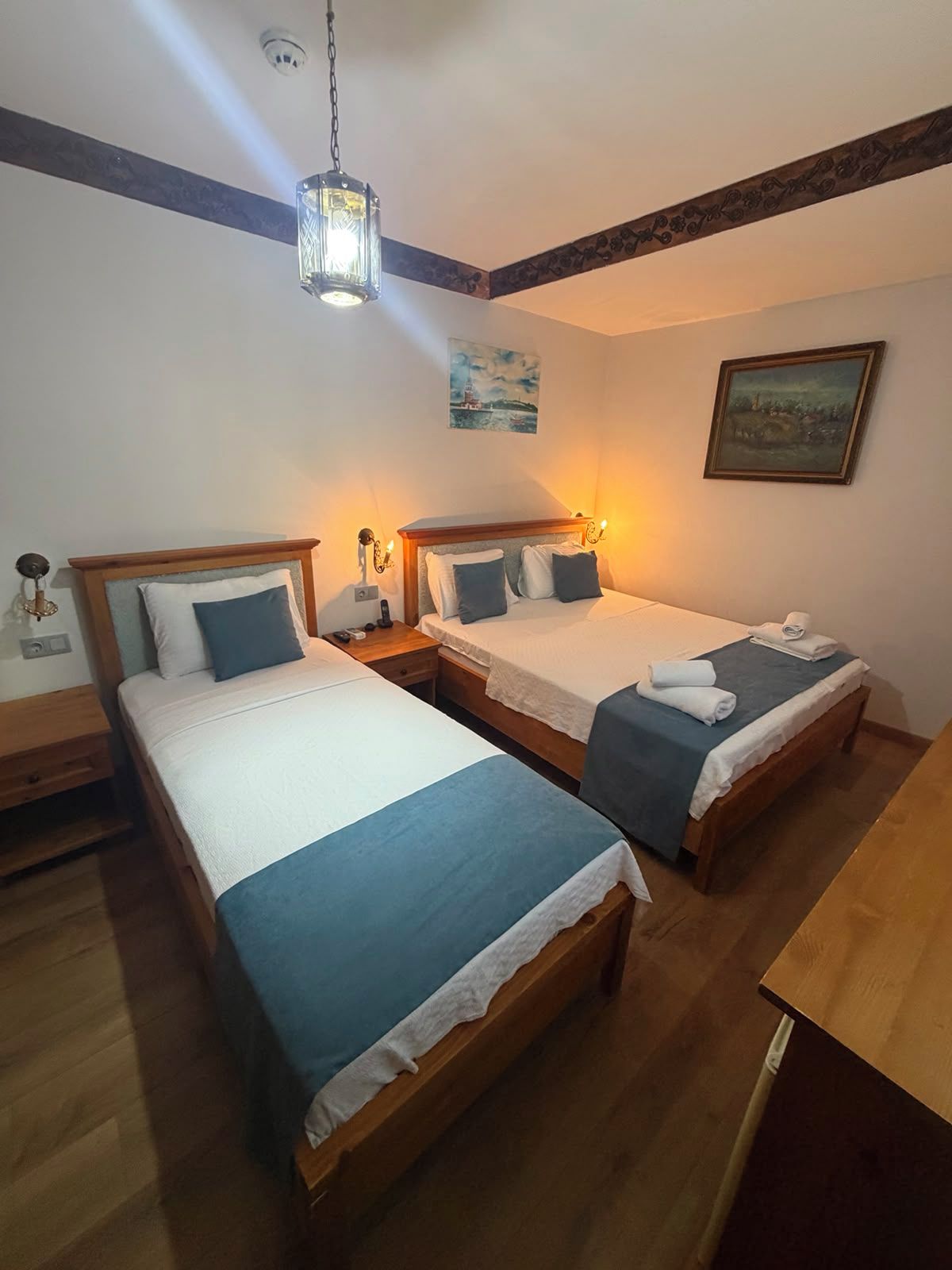A hotel room with two single beds, wooden headboards, bedside tables, a hanging light fixture, and a painting on the wall.