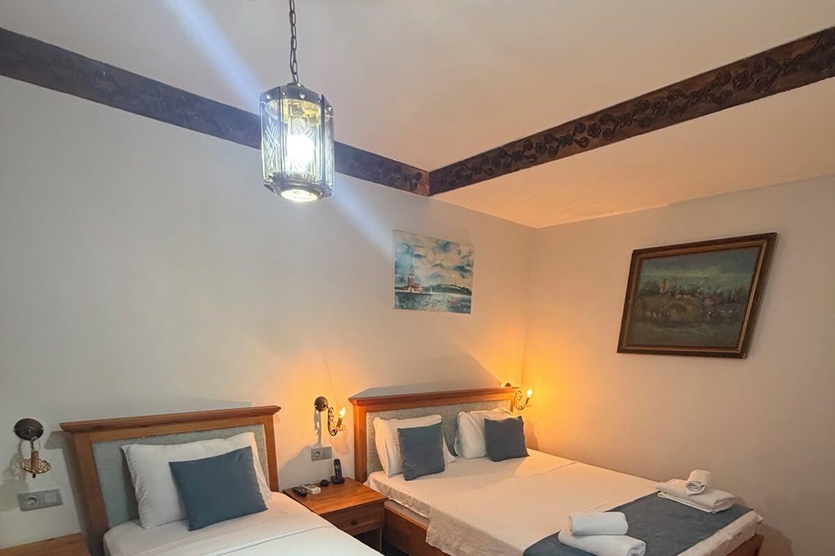 A hotel room with two single beds, wooden headboards, bedside tables, a hanging light fixture, and a painting on the wall.