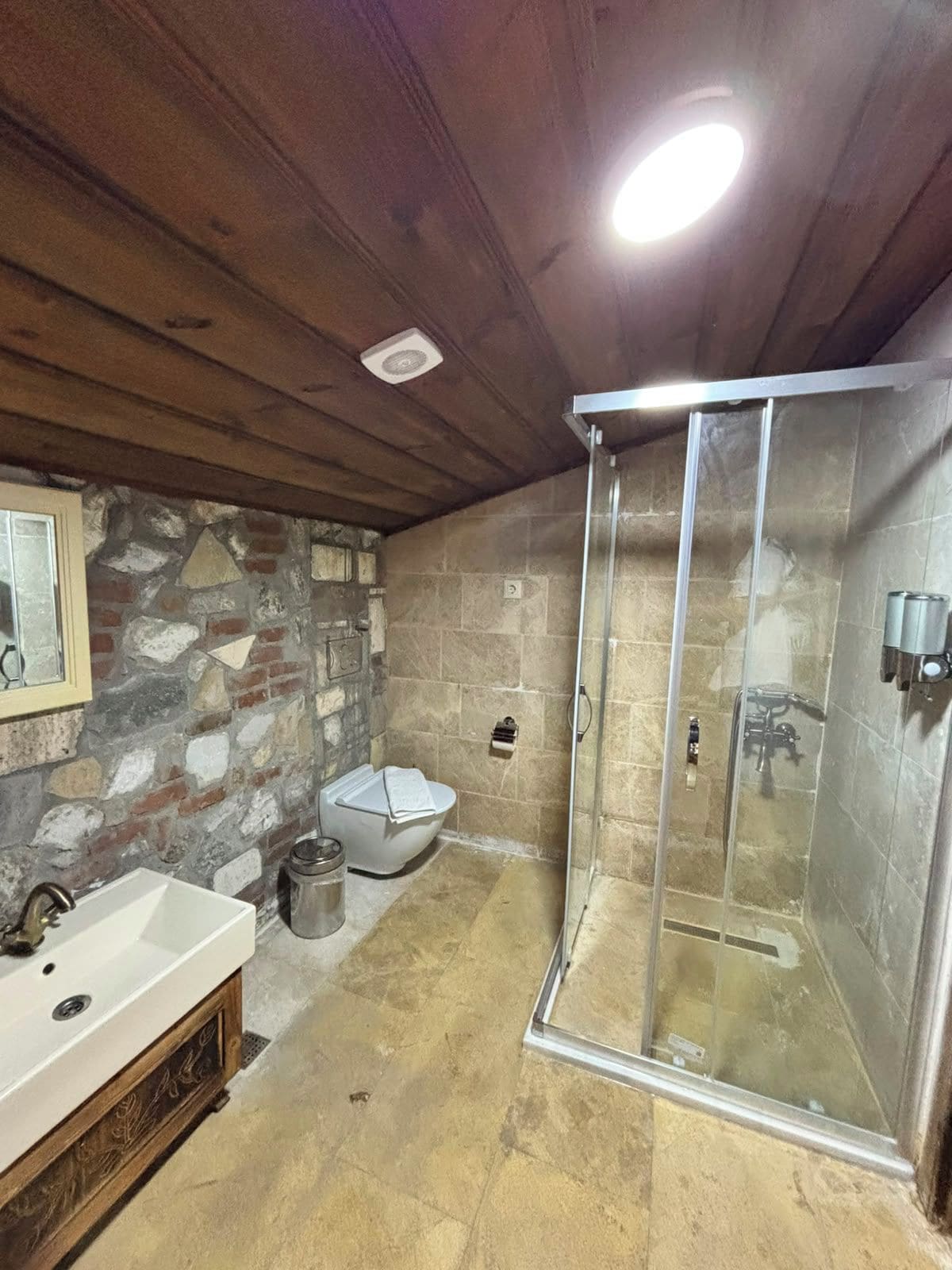 Un baño con pared de piedra, lavamanos blanco, inodoro y ducha con cerramiento de vidrio.