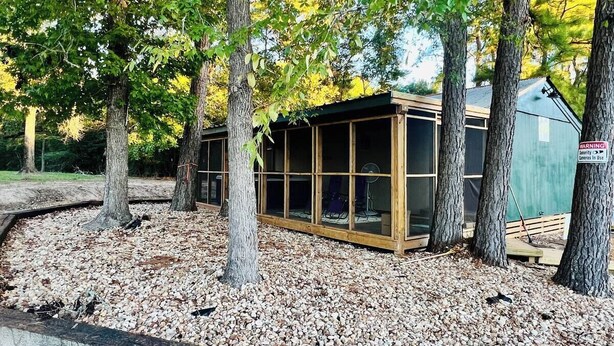 A small, enclosed structure with a glass front, surrounded by trees and gravel. - UPSCALE EAST TEXAS CABIN, PRIVATE HOT TUB, WIFI, ROKU, OLD MILL VILLAGE KENNARD (Kennard)