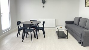 Un salon moderne avec un canapé gris, une table de salle à manger ronde entourée de quatre chaises, une petite table basse, une horloge murale et un radiateur.