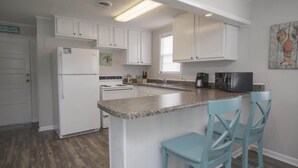 Una cocina con armarios blancos, encimera de granito, refrigerador, estufa, pileta y dos sillas azules.