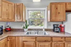 A kitchen with wooden cabinets, a granite countertop, a double sink, a coffee maker, and a view of trees through a window. - Bearden Bungalow - 10 Min. to Downtown and West Knoxville (Knoxville)