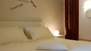 Una cama bien tendida con ropa de cama y almohadas blancas, una lámpara de noche y una puerta con un panel de vidrio decorativo.