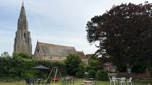 A grassy outdoor area with picnic tables, chairs, and a swing set. A church with a tall spire is visible in the background. - The Queens Road Hotel (Shanklin)