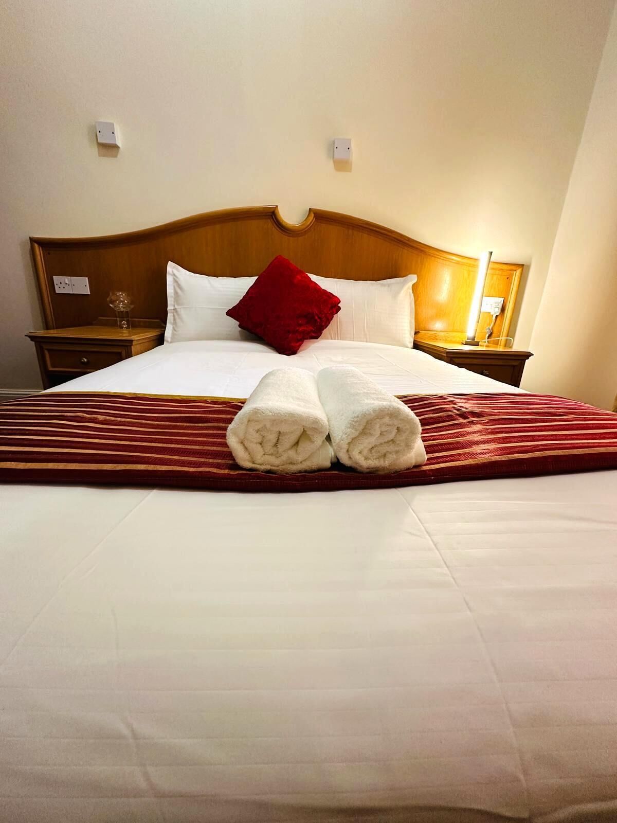 Una cama bien hecha con un almohadón rojo, dos toallas enrolladas y una manta a rayas.