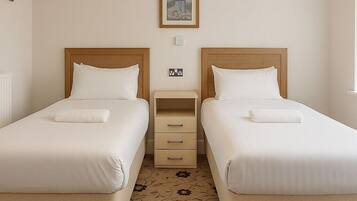 A hotel room with two single beds, a small bedside table, a lamp, a framed picture on the wall, and a floral carpet.
