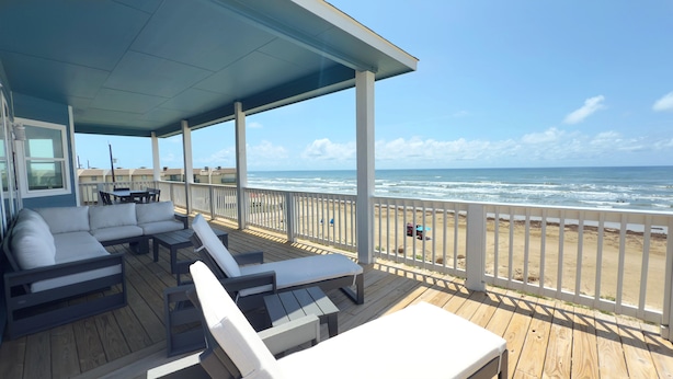 A covered outdoor deck with a sofa and chairs, overlooking a beach with waves.