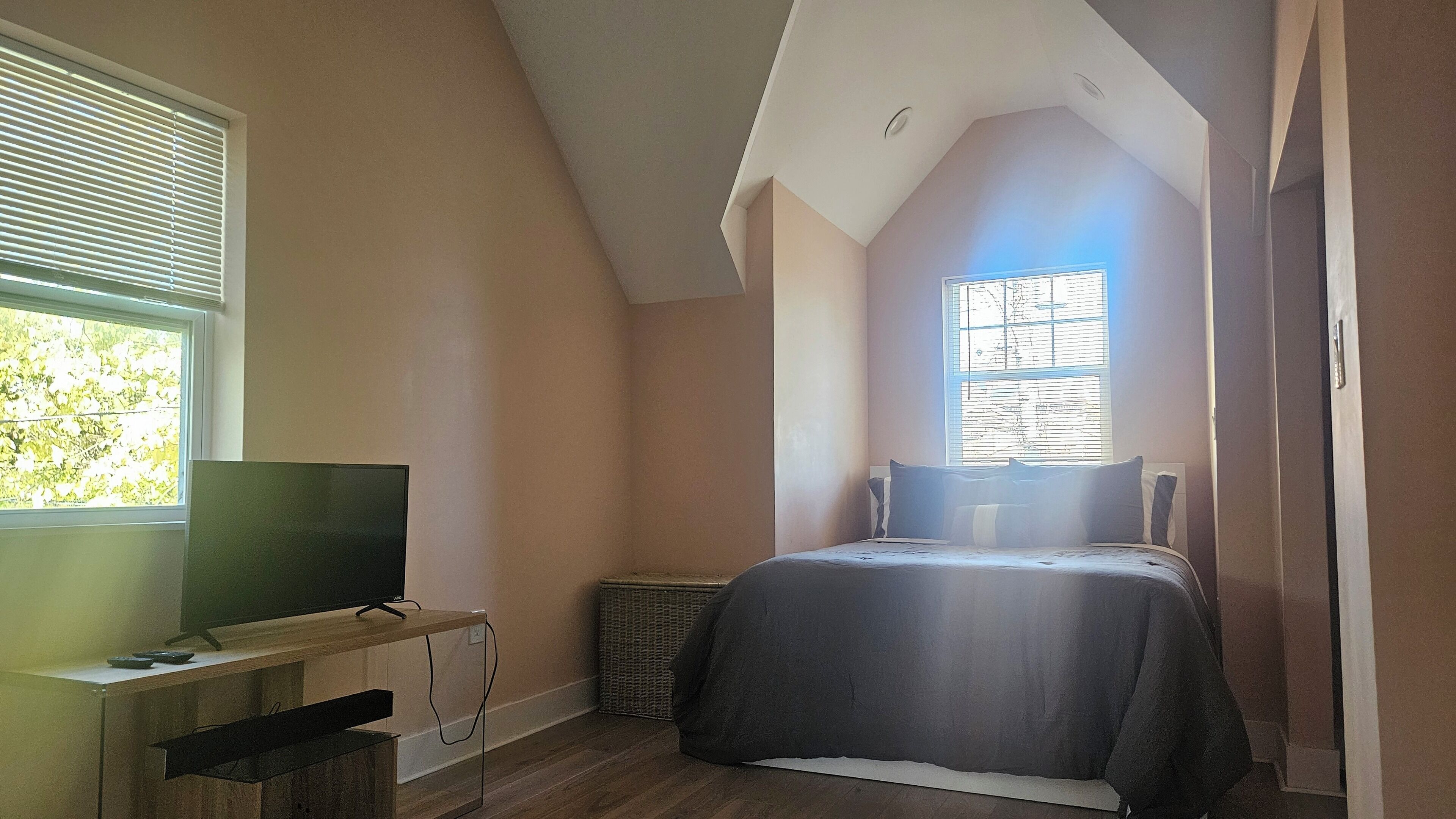 Une chambre à coucher avec un lit, un téléviseur posé sur un meuble en bois, une fenêtre avec des stores et un plafond doté d’un puits de lumière.
