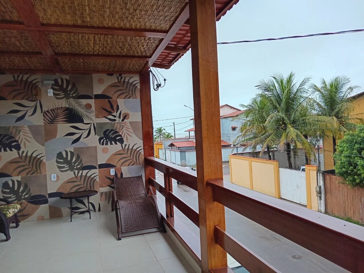 Un espacio exterior techado con un muro decorado, baranda de madera y vista a una calle residencial con palmeras.