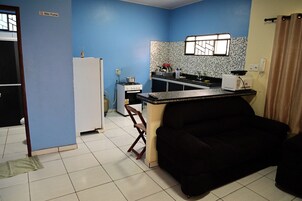 Cocina compacta con refrigerador, estufa y lavamanos. Un sofá negro se encuentra en la sala.