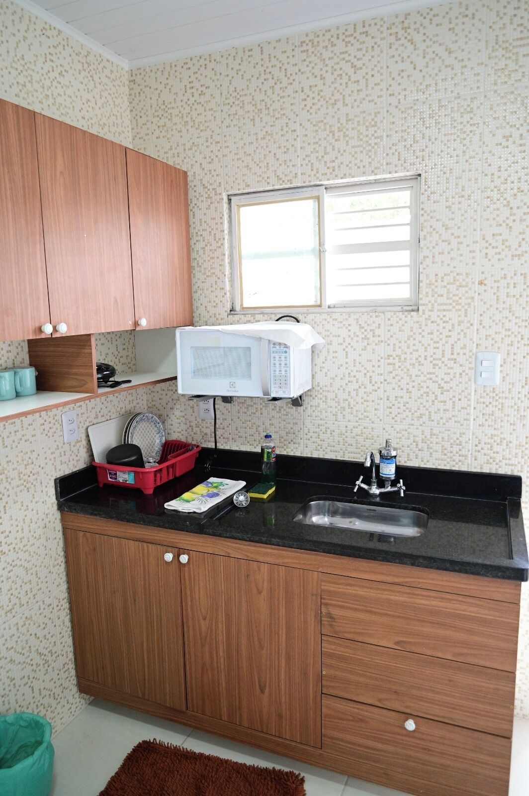 Una cocina con armarios de madera, encimera negra, microondas, pileta y ventana.