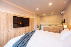 Una habitación de hotel moderna con una cama grande, un televisor de pantalla plana montado en la pared, una kitchenette pequeña y un equipo de aire acondicionado instalado en el techo.
