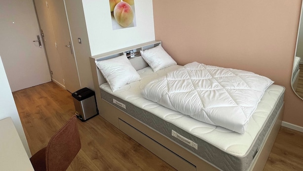Une chambre bien rangée, avec un lit, une petite poubelle et une porte.