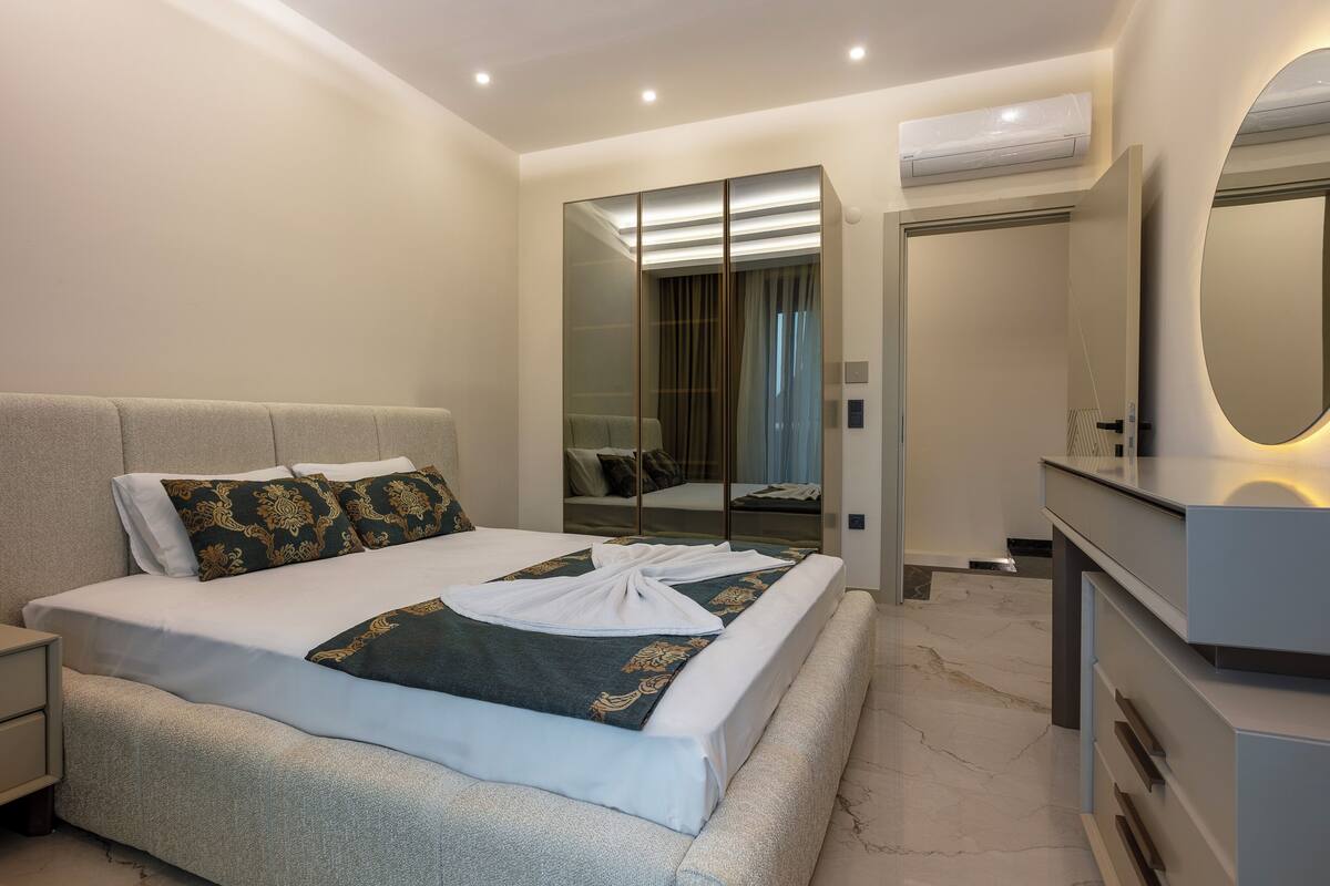 A modern hotel room with a large bed, a bedside table, a wardrobe, a mirror, and a built-in air conditioning unit.