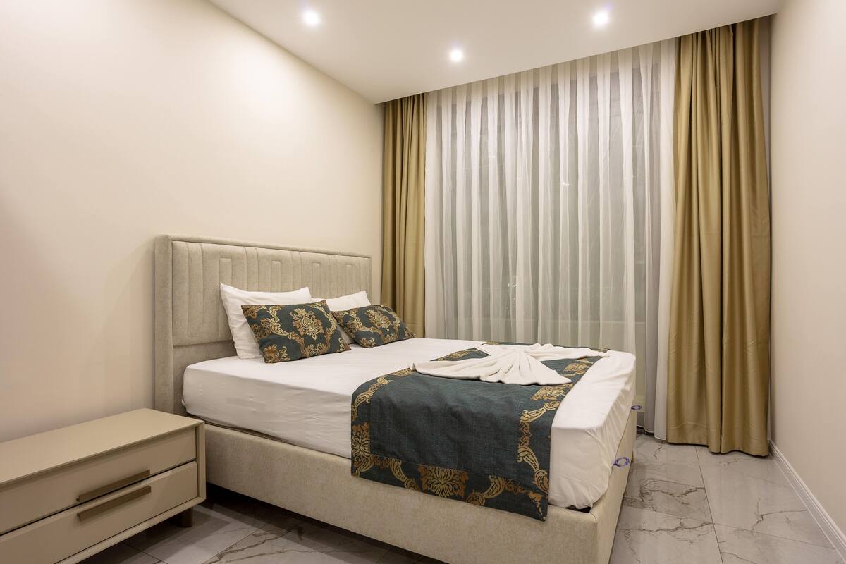 A neatly made bed with a decorative throw, a bedside table with drawers, and a large window with sheer curtains.