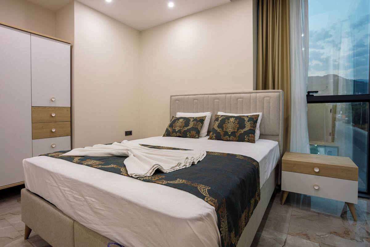 A neatly made bed with a white and dark patterned bedspread, a wooden nightstand with drawers, and a view of the outdoors through a window with curtains.
