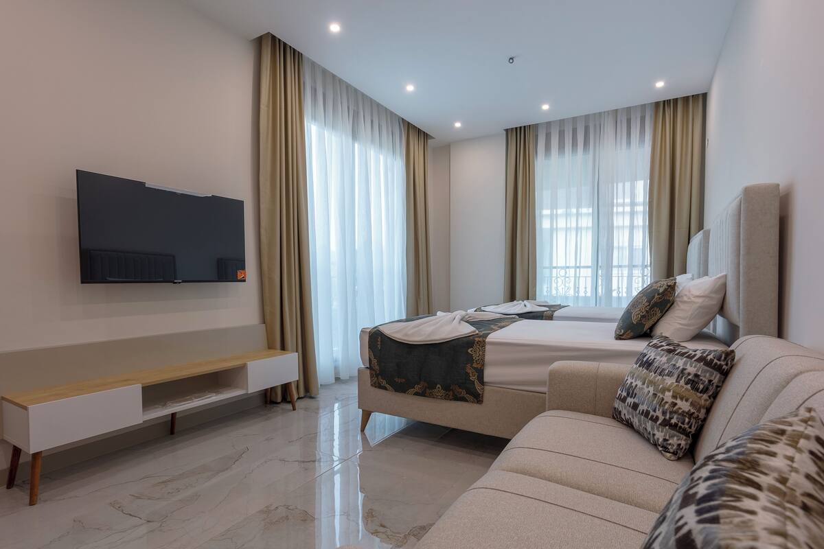 A modern hotel room with a bed, a sofa, a TV mounted on the wall, and a large window with curtains.