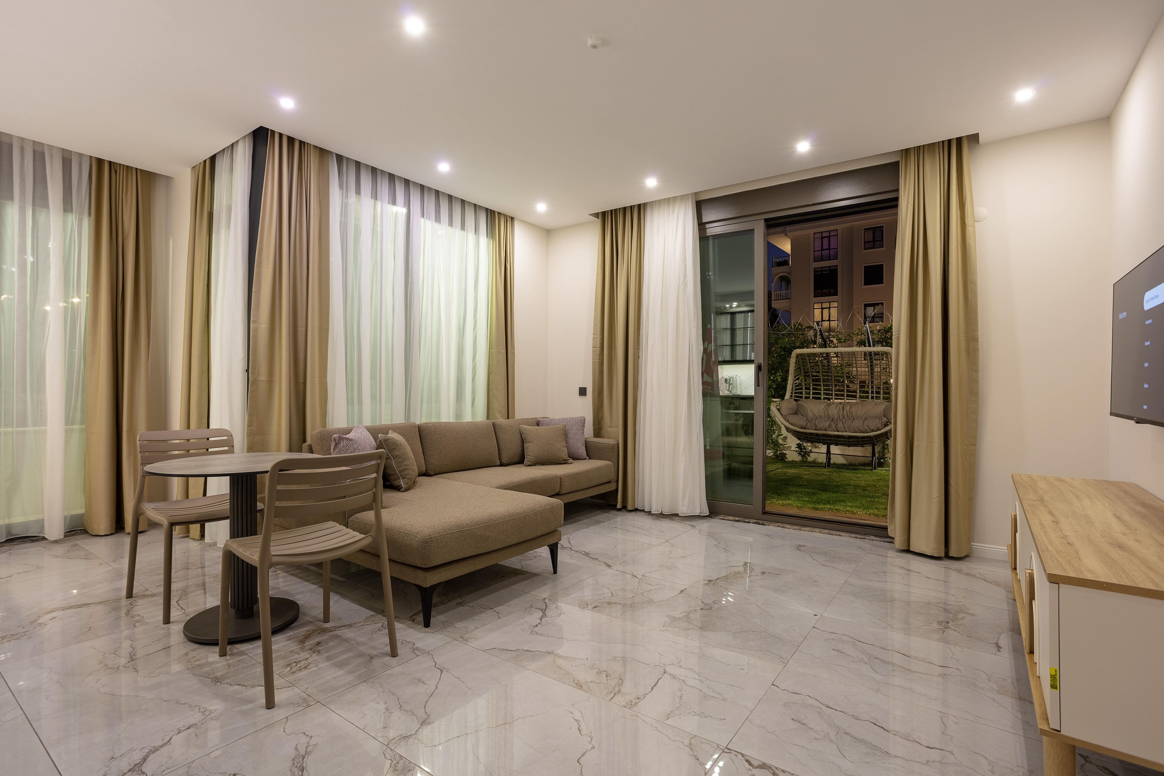 A modern living room with a sectional sofa, a round table, and chairs, a TV, and a glass door leading to a balcony with a chair outside.