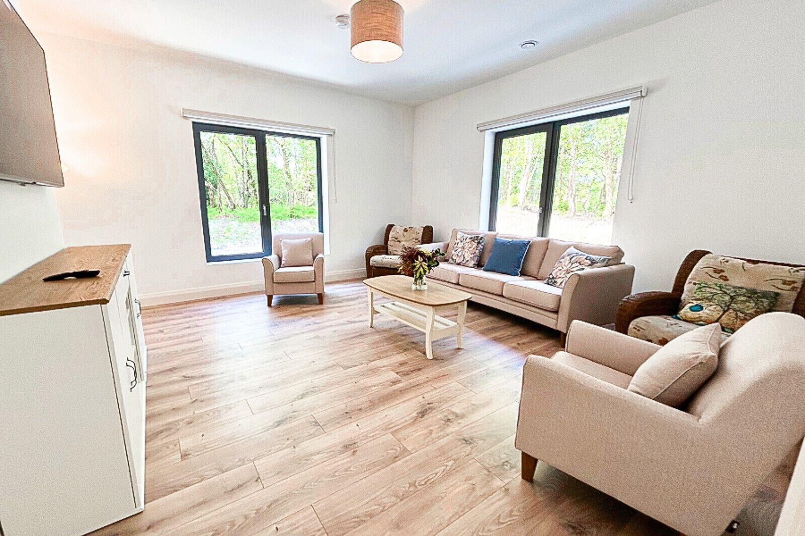 A spacious living room with a sofa set, a coffee table, and a TV on a stand. There are two windows providing natural light and a view of greenery outside.