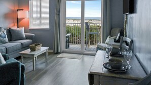 Un salón con sofá, una mesa de centro y una mesa de comedor para dos personas. Se ve la playa a través de puertas de vidrio.