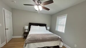 Une chambre à coucher avec un lit, un ventilateur de plafond, une table de chevet, une lampe et une fenêtre avec des stores.