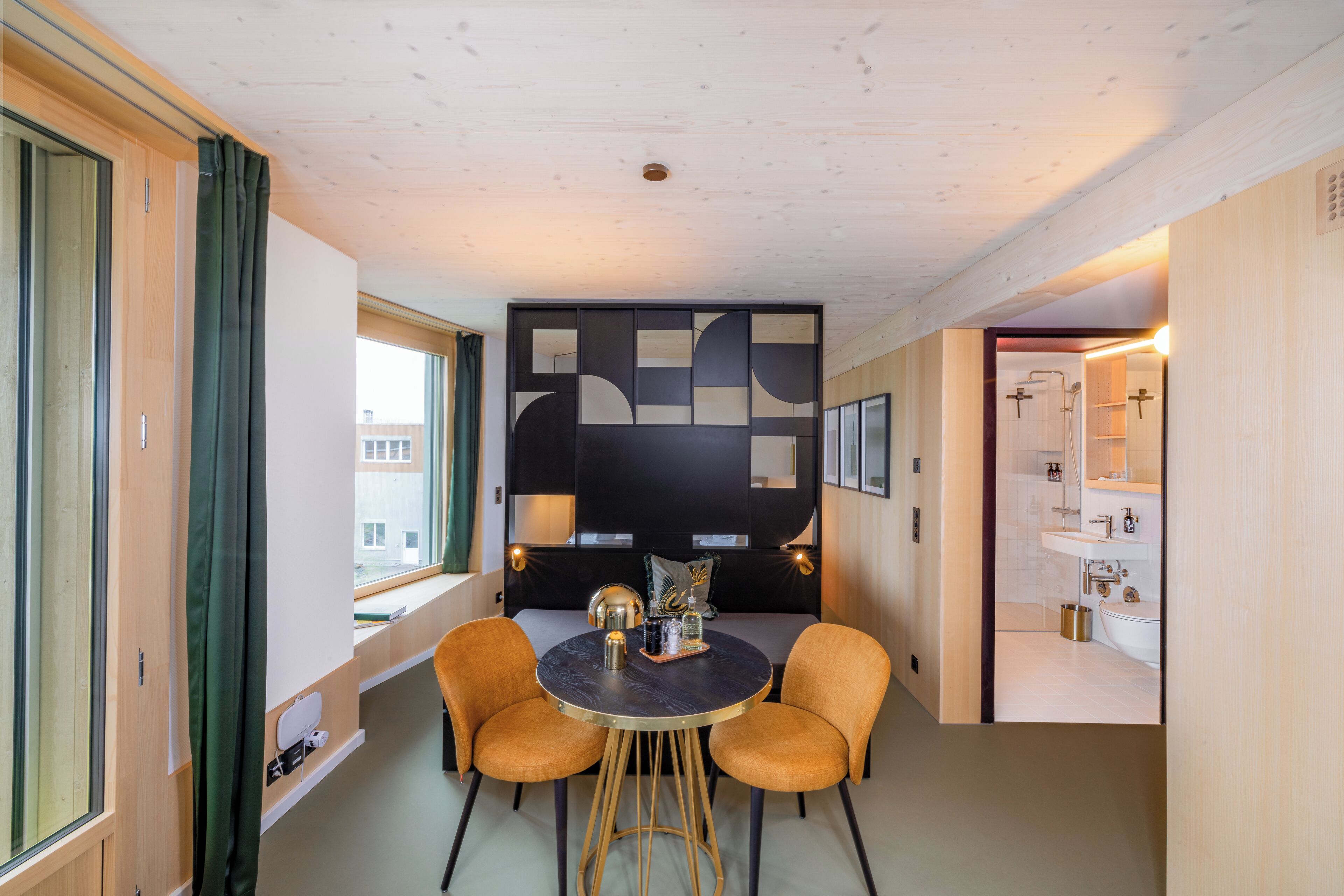 Een moderne kamer met een ronde tafel, twee stoelen, een decoratieve wand, en een zichtbare badkamer door een geopende deur.