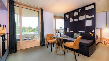 A modern hotel room with a balcony, a round dining table, two chairs, a sofa, and a decorative wall panel.