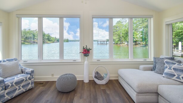 Un ampio soggiorno con ampie finestre vista lago, una ventola a soffitto e varie opzioni di seduta, tra cui un divano decorato e un pouf.