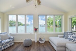 A spacious living room with large windows overlooking a lake, a ceiling fan, and a mix of seating options including a patterned sofa and a bean bag.