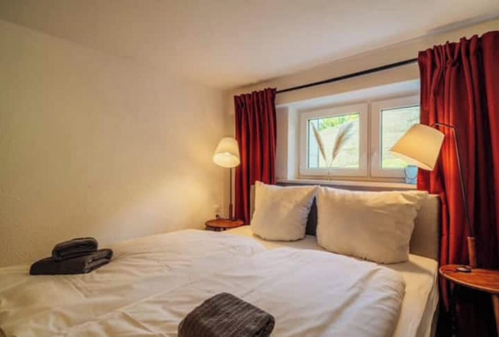 Una habitación de hotel con una cama, dos lámparas de noche, un ventanal con cortinas y una mesita con lámpara.