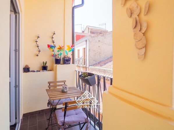 Un balcón con mesa y sillas de madera, plantas en macetas y vista de edificios al fondo.