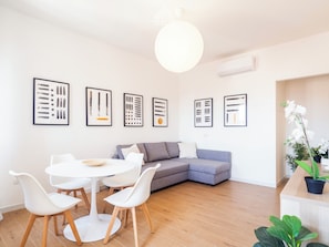 Ein modernes Wohnzimmer mit grauem Sofa, weißen Sesseln, einem runden Tisch und abstrakten Kunstwerken in Rahmen an der Wand.