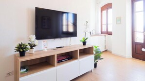 A living room with a flat-screen TV, wooden entertainment unit, potted plants, and a view of a window with a door. - Sa Perda e Su Entu - Morada (Iglesias)