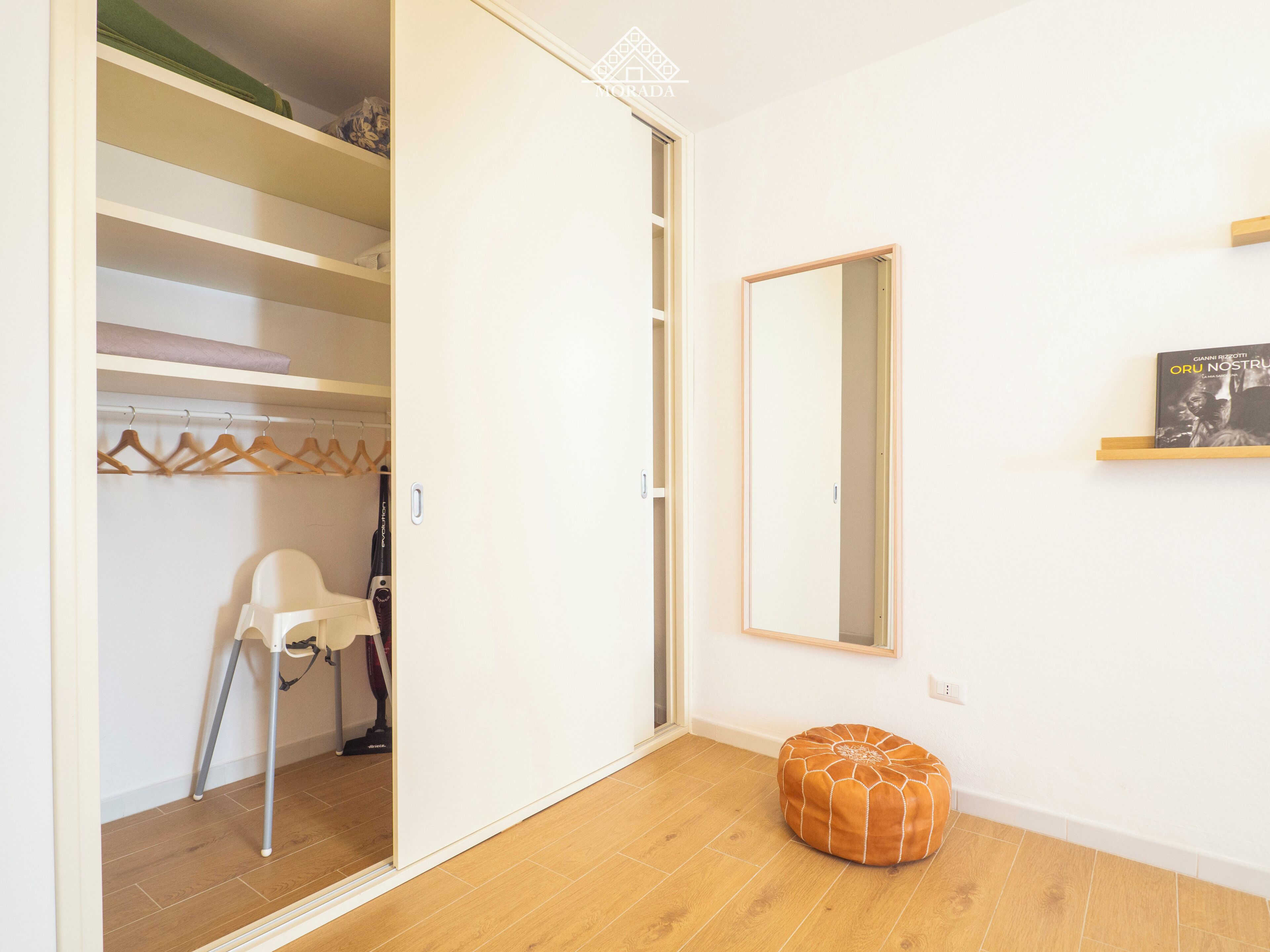 Een kamer met een houten vloer, een witte kledingkast met planken, een hangstoel, een stofzuiger, een rond poefje en een lijst met een foto aan de muur.