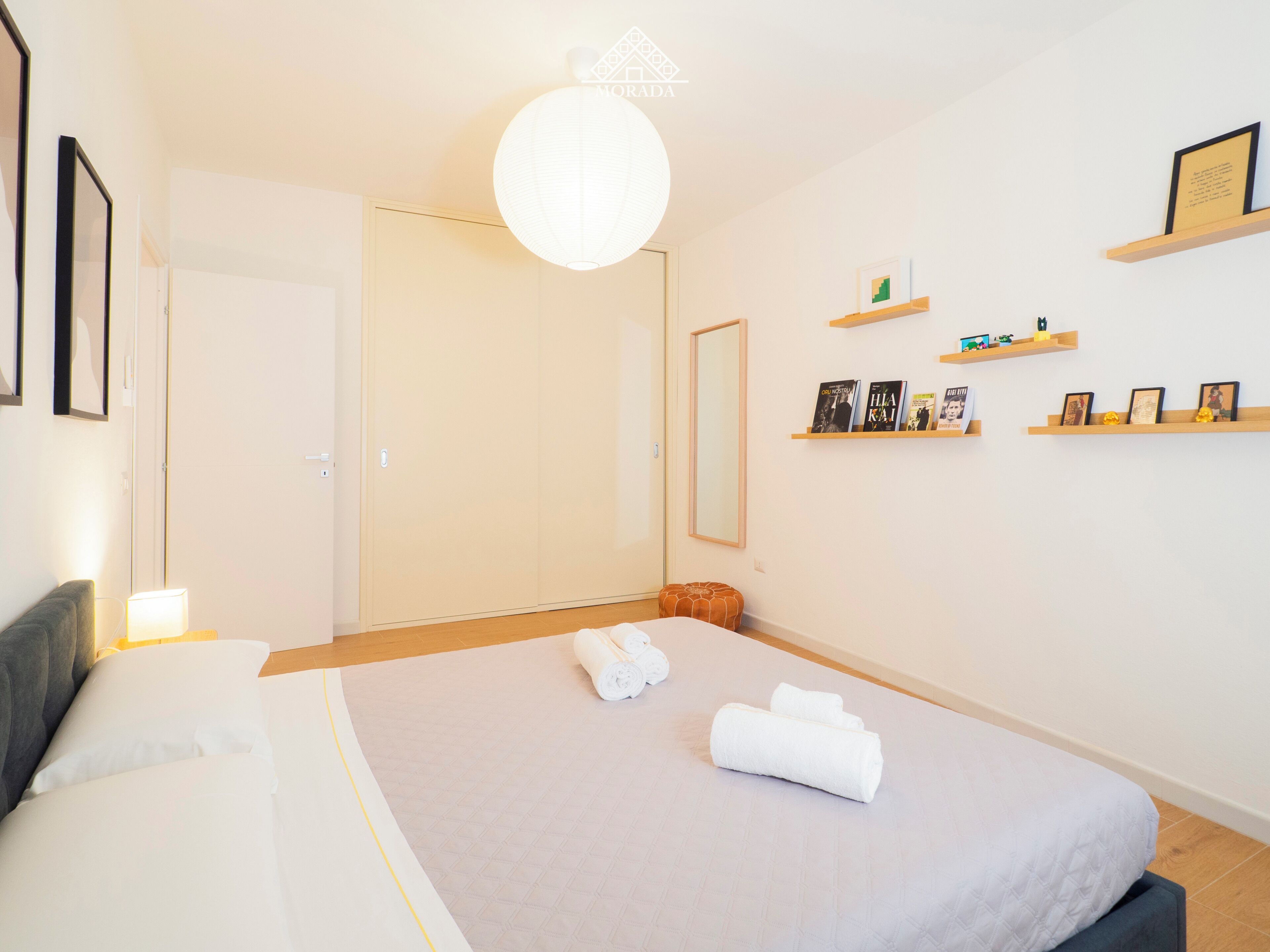 A bedroom with a bed, a bedside table, a lamp, a wardrobe, a mirror, and shelves with books and pictures.