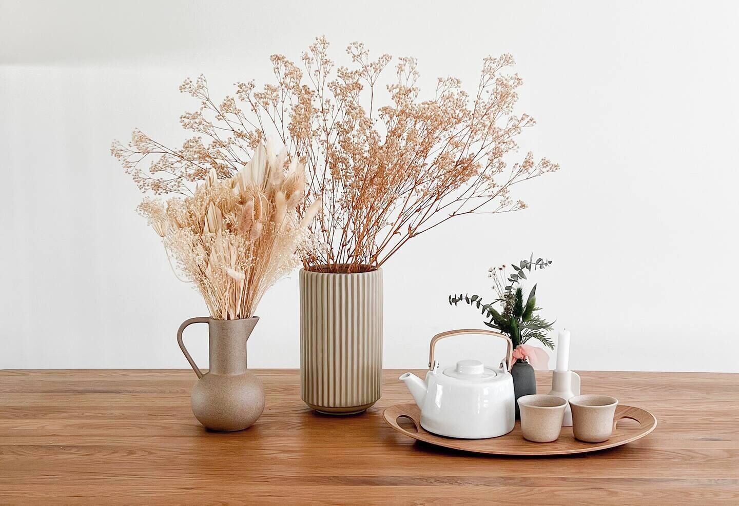 Une table en bois, sur laquelle se trouvent une théière blanche, un vase beige contenant des fleurs séchées et une petite plante en pot.