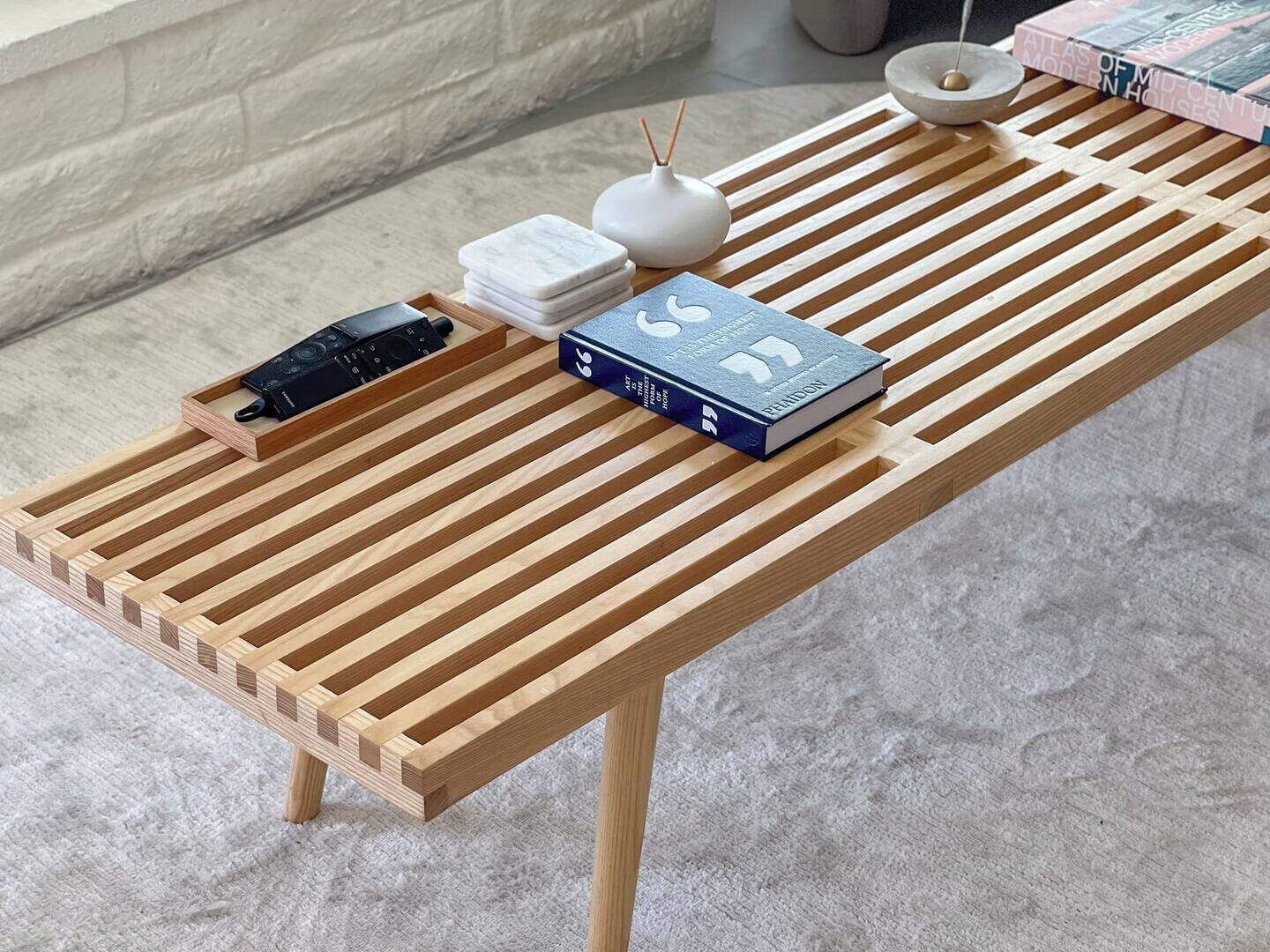 Une table en bois au design à lattes, un livre intitulé « Atlas des maisons de style moderniste du milieu du XXe siècle », un vase blanc et un petit objet noir posé sur la table.