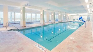 En indendørs swimmingpool med krystalklart vand, flisegulv og udsigt til byen gennem store vinduer.